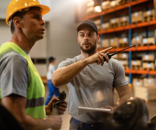 Young dispatcher communicating about delivery schedule with warehouse worker in factory warehouse.